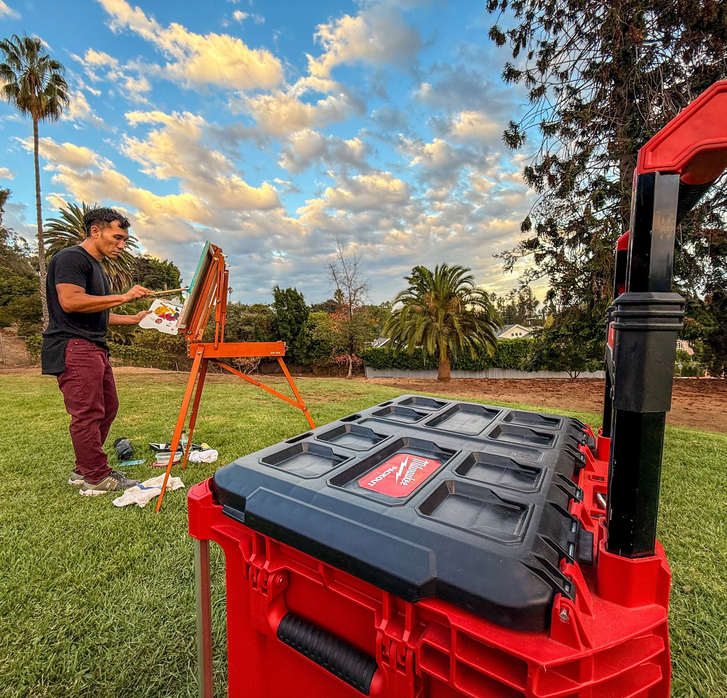 Painter at an easel beside a red Milwaukee tool case in a park